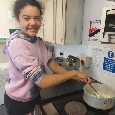 A child in a pink jumper stands at a hob cooking some onions in a saucepan. She smiles at the camera.
